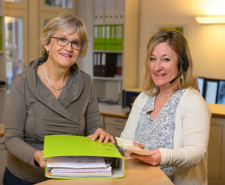 Zwei lächelnde Frauen sitzen mit einem grünen Ordner am Tisch